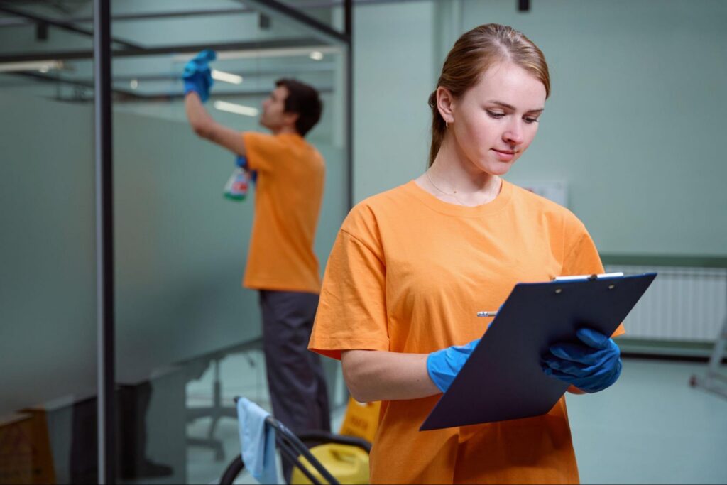 Cleaning supervisor reviewing an inspection checklist while a crew member wipes office glass.